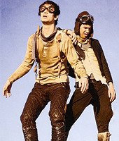 Ryan Ross and Brendon Urie in vintage pilot outfits standing together against a blue sky.